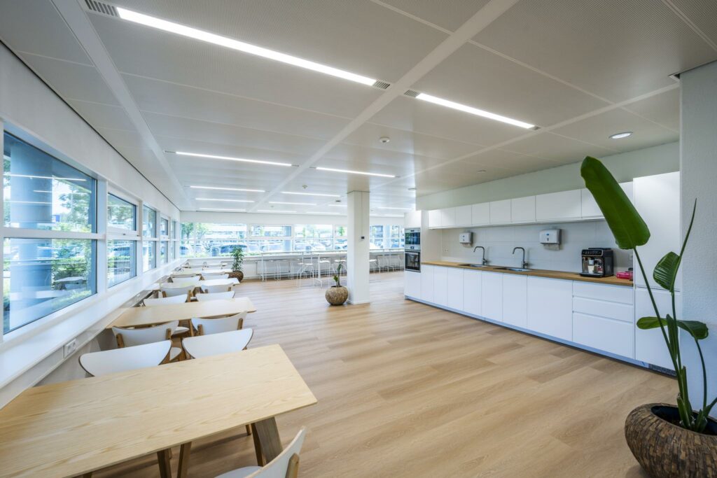 Moderne, lichte bedrijfskantine aan de Burgemeester Stramanweg met houten vloeren, witte keuken en uitzicht op parkeerterrein.